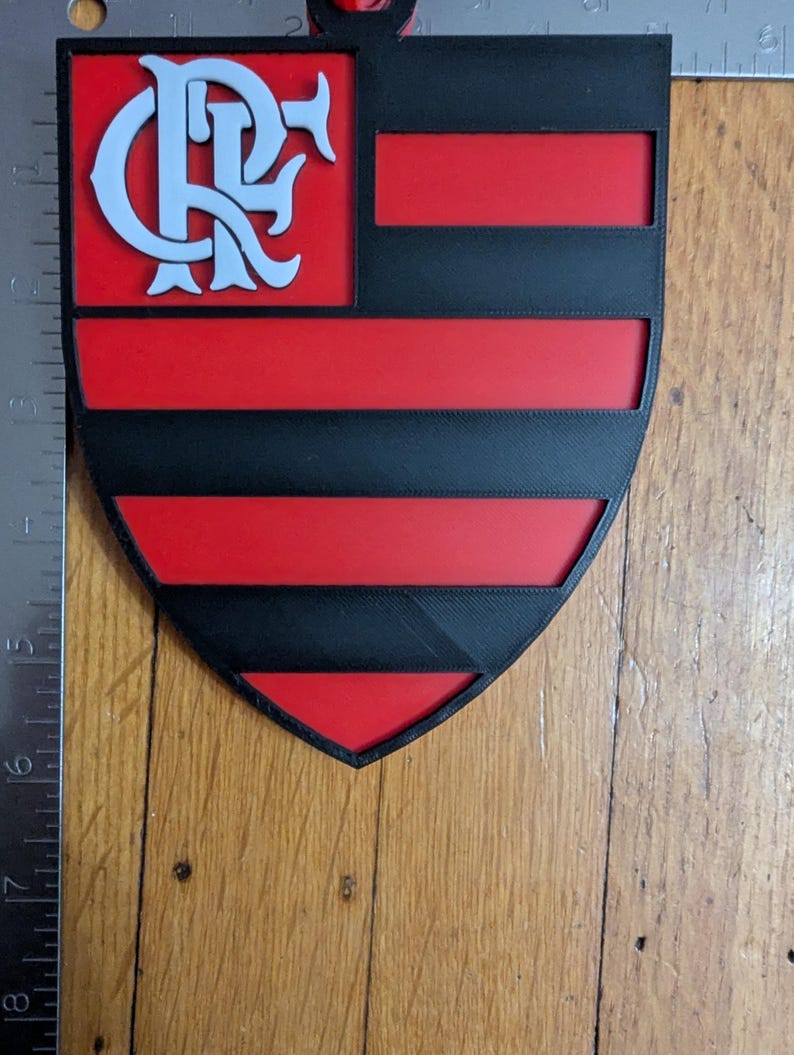 May include: A shield-shaped emblem featuring the Flamengo logo in white on a black and red background. The design includes the team's initials and horizontal stripes, with a small loop at the top for hanging.