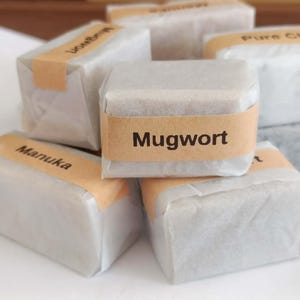 May include: Several wrapped soap bars with tan labels. The central bar has the word "Mugwort" printed on it in black. Other labels read "Manuka", "Lion's Mane", and "Pure Chia". The bars are wrapped in white paper.