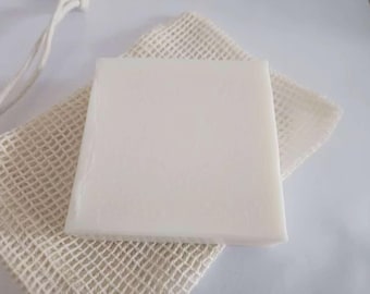 May include: A square, off-white bar of soap sits on a beige, mesh soap saver. A white string is attached to the soap saver. The soap has a smooth surface and a slightly translucent appearance. The soap saver has a woven texture.