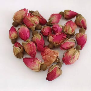 May include: A heart shape made from dried rose buds. The buds are a mix of pink, red, and tan, with a natural, organic appearance. The arrangement is set against a plain white background.