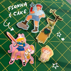 May include: A collection of colorful stickers featuring characters from the animated series "Fionna & Cake." The stickers depict various characters in action poses, with vibrant colors and detailed designs. The stickers are on a green cutting mat.