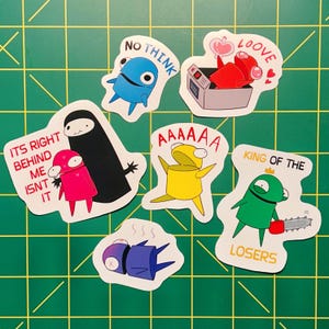 May include: A selection of seven cartoon stickers in various colours. The stickers feature text such as "NO THINK", "LOOVE", "AAAAAA", and "KING OF THE LOSERS". The stickers are displayed on a green cutting mat.