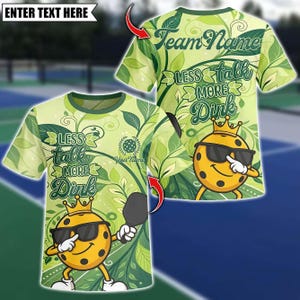 Pode incluir: Uma camiseta verde clara com um padrão de folhas e o texto "LESS talk MORE Dink". A camiseta apresenta um personagem de pickleball de desenho animado usando óculos de sol e uma coroa, segurando uma raquete. As palavras "Team Name" e "Your Name" também são visíveis.
