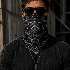 May include: A black neck gaiter featuring a white, intricate, symmetrical fractal pattern that resembles a skull. The fabric covers the lower face, and the design includes a small circular emblem at the top centre, paired with black sunglasses.