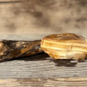 May include: Two natural stones rest on a wooden surface. One is a long, smooth, brown and black stone. The other is a layered, tan and cream-colored stone with a rough texture. The stones are illuminated by sunlight.