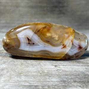 May include: Polished agate stone with a smooth, rounded shape. The stone features bands of white, brown, and amber colors, creating a natural, layered pattern. The surface is glossy, reflecting light.