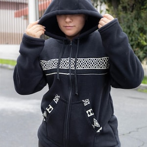 May include: A black hooded sweatshirt with a full-length zipper and white geometric patterns across the chest and on the pockets. The hood is up, and the sweatshirt appears to be made of a soft, warm material. The person is wearing black leggings.