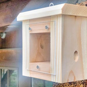 May include: A wooden birdhouse with a light-colored finish, featuring a rectangular opening with a clear plastic panel secured by small screws. The birdhouse has a small circular entrance hole and a sloped roof. It is mounted on a textured surface.