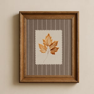 May include: A framed wall art piece featuring a dried, golden-brown maple leaf centred on a cream-coloured fabric patch with pinked edges. The patch is mounted on a dark taupe background with thin, vertical white pinstripes in a wooden frame.