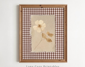 May include: A framed wall art piece featuring a single pressed cream-colored flower with leaves on a textured beige paper background with deckled edges, set against a brown and white gingham pattern, with the text Lana Cozy Printables below.