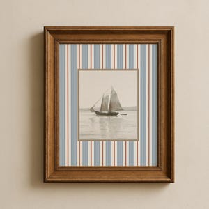 Puede incluir: Un cuadro enmarcado con un velero en el agua, en tonos sepia. El marco es marrón y el fondo tiene rayas verticales en azul, rojo y crema. La obra de arte es cuadrada.