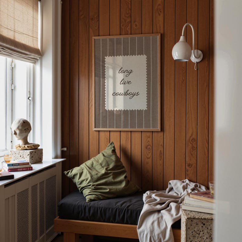 May include: A framed print with the words "long live cowboys" in a room with wood paneling. The print is centered on the wall above a black cushion and green pillow. A white lamp is mounted on the wall. A window with a beige shade is on the left.