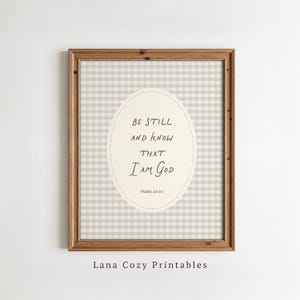 May include: A framed wall art print featuring a light beige and white gingham pattern background. In the center, an oval shape contains the black text: Be Still and know that I am God, Psalm 46:10. The frame is made of medium-toned brown wood.