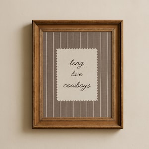 May include: A framed print with a brown wooden frame. The print features a gray and white striped background with a cream-colored, scalloped-edged rectangle in the center. The text "long live cowboys" is written in a cursive font.