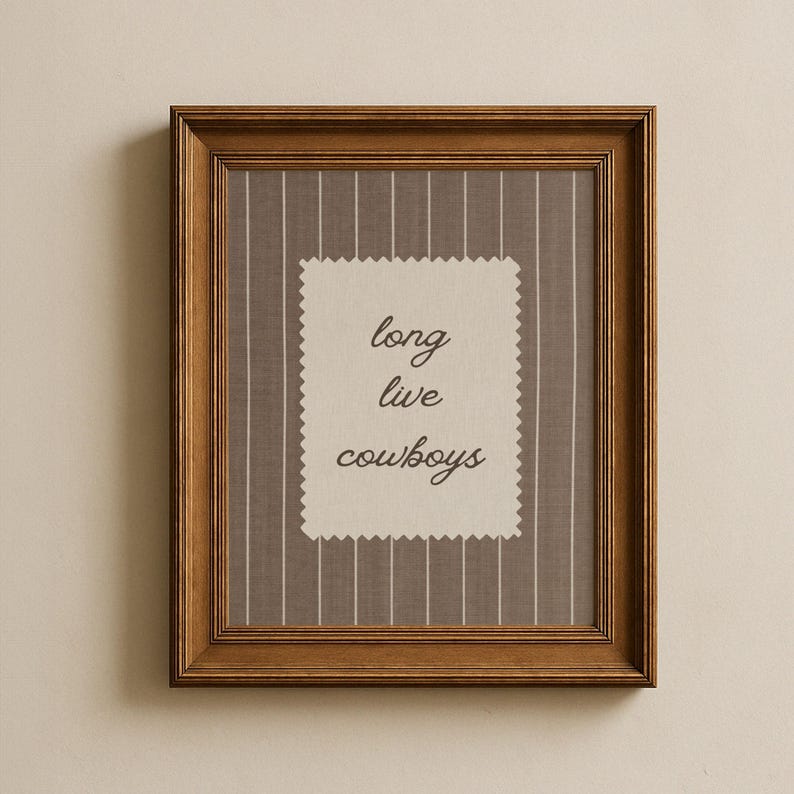 May include: A framed print with a brown wooden frame. The print features a gray and white striped background with a cream-colored, scalloped-edged rectangle in the center. The text "long live cowboys" is written in a cursive font.