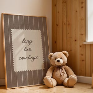 May include: A framed artwork with the text "long live cowboys" in a room with a teddy bear. The artwork has a gray and white striped background with a cream-colored square in the center. A large, brown teddy bear with a bow sits next to the artwork.