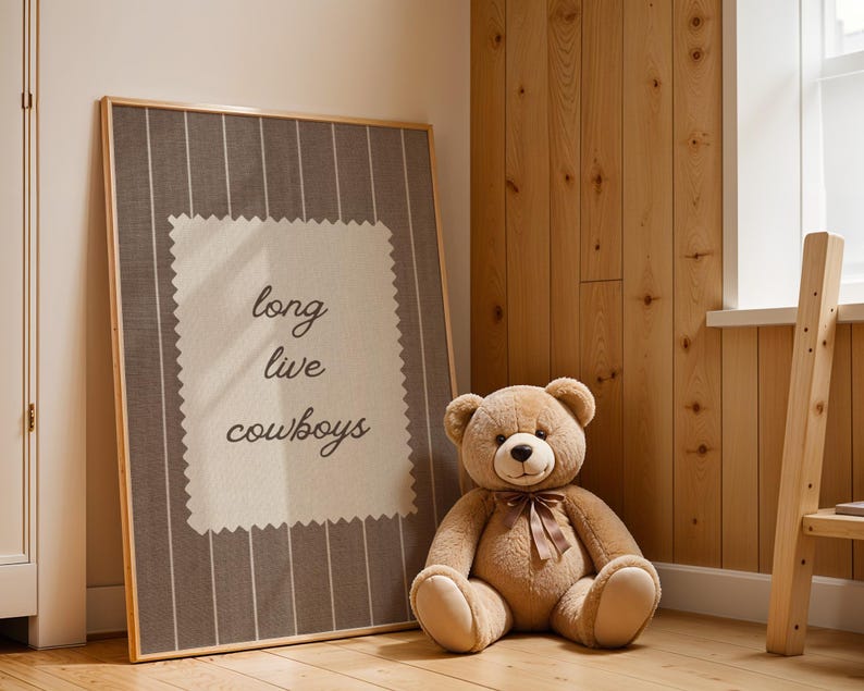 May include: A framed artwork with the text "long live cowboys" in a room with a teddy bear. The artwork has a gray and white striped background with a cream-colored square in the center. A large, brown teddy bear with a bow sits next to the artwork.
