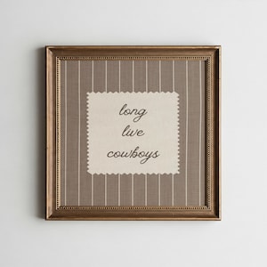 May include: A framed artwork with a gold-colored frame. The artwork features a beige background with vertical white stripes. A cream-colored, scalloped-edge patch in the center displays the words "long live cowboys" in black script.