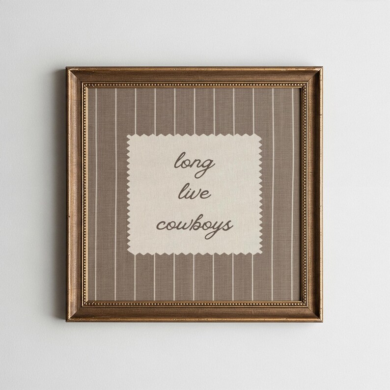 May include: A framed artwork with a gold-colored frame. The artwork features a beige background with vertical white stripes. A cream-colored, scalloped-edge patch in the center displays the words "long live cowboys" in black script.