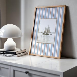 May include: A white mushroom-shaped table lamp sits atop a stack of books labeled "TOM FORD" and "TOM FORD GENTLE LIVING." A framed picture of a sailboat is propped against a wall. The frame has a wood grain finish and a blue and white striped background.