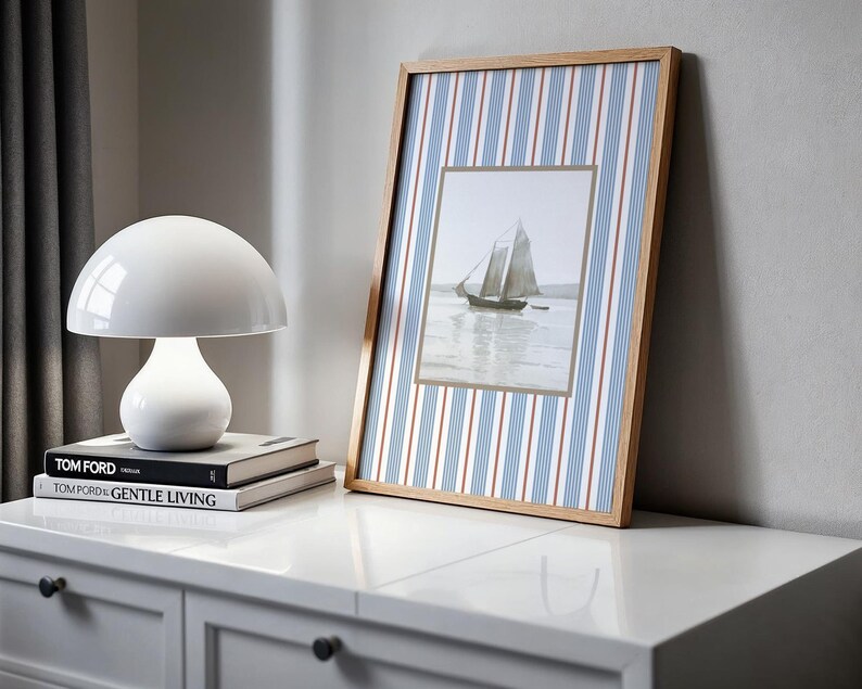 May include: A white mushroom-shaped table lamp sits atop a stack of books labeled "TOM FORD" and "TOM FORD GENTLE LIVING." A framed picture of a sailboat is propped against a wall. The frame has a wood grain finish and a blue and white striped background.