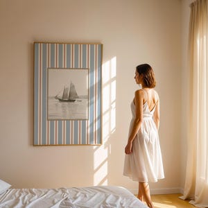 May include: A framed nautical print of a sailboat on the water hangs on a wall. The frame has a blue, white, and red striped border. A woman in a white dress stands in front of the artwork in a sunlit room.