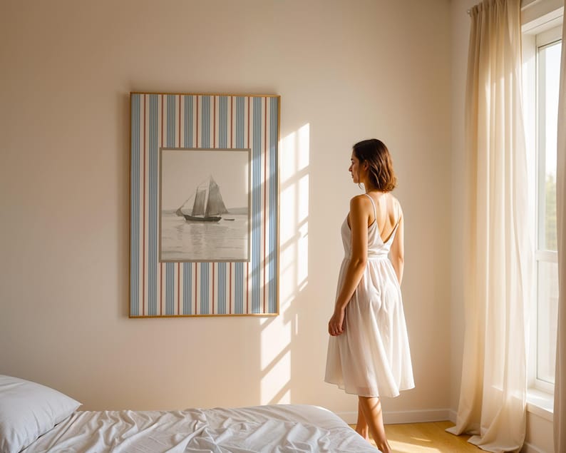 May include: A framed nautical print of a sailboat on the water hangs on a wall. The frame has a blue, white, and red striped border. A woman in a white dress stands in front of the artwork in a sunlit room.