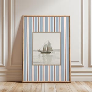 May include: Framed nautical art print featuring a sailboat on the water. The artwork is set within a brown frame and a striped background of blue, red, and white vertical lines. The print is displayed against a white wall.