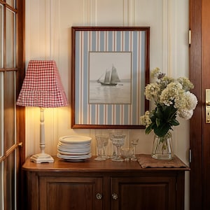 May include: A wooden cabinet with a lamp, framed sailboat art, and glassware. The lamp has a red and white checkered shade. The artwork features a sailboat on the water. A vase of white flowers sits on the cabinet.