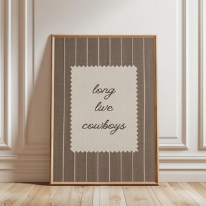 May include: Framed artwork with a brown and white striped background. A cream-colored rectangle in the center features the words "long live cowboys" in a cursive font. The frame is a light brown color.