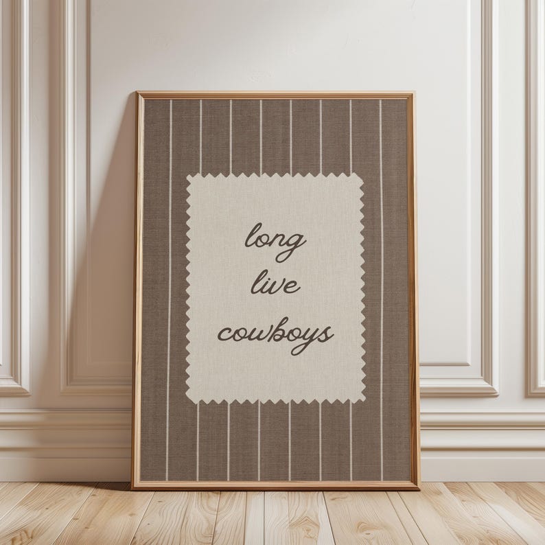 May include: Framed artwork with a brown and white striped background. A cream-colored rectangle in the center features the words "long live cowboys" in a cursive font. The frame is a light brown color.