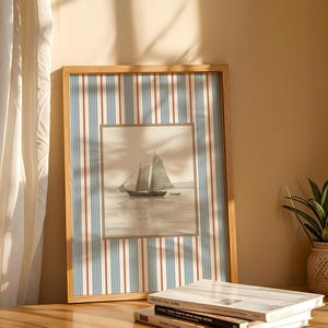 May include: A framed nautical print featuring a sailboat on the water. The frame has a striped pattern in blue, red, and white. The print is displayed on a wooden surface, next to a small potted plant and books.