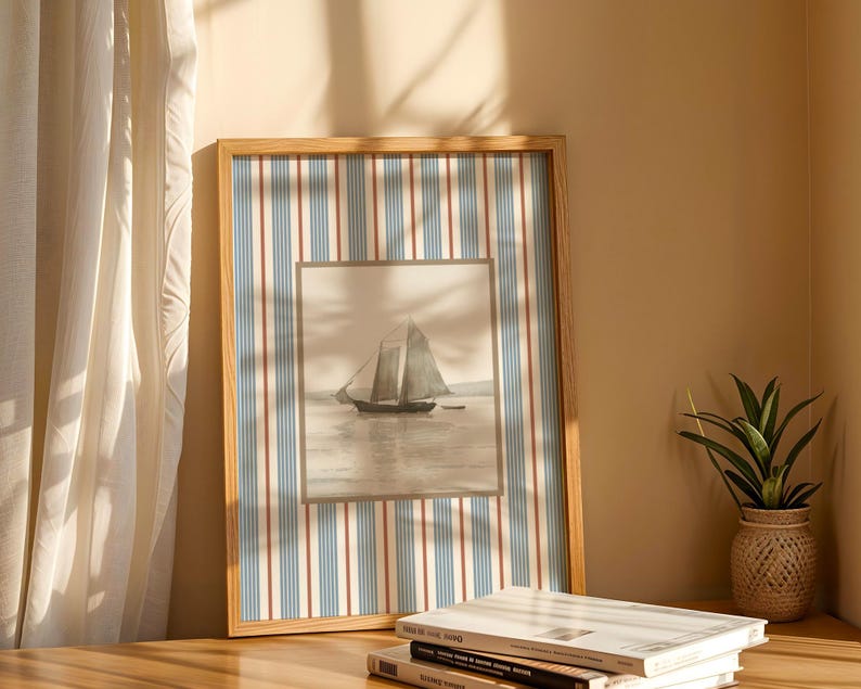 May include: A framed nautical print featuring a sailboat on the water. The frame has a striped pattern in blue, red, and white. The print is displayed on a wooden surface, next to a small potted plant and books.