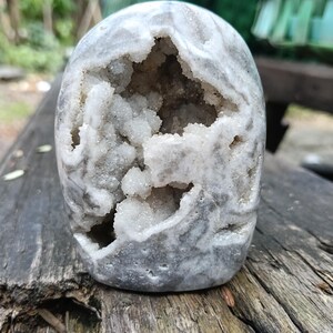 Puede incluir: Una piedra de geoda gris y blanca con un centro hueco lleno de cristales brillantes. La piedra tiene forma redondeada y un patrón veteado. La geoda se exhibe sobre una superficie de madera envejecida.