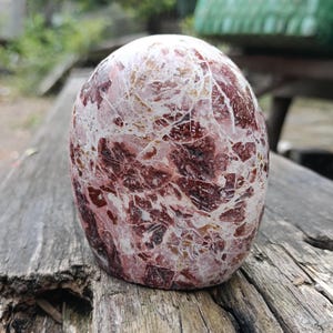 May include: A polished, oval-shaped stone with a marbled appearance. The stone features a mix of deep red, white, and brown colors, with a network of fine white veins running throughout. The stone is displayed on a weathered wooden surface.