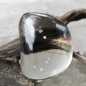 Op de afbeelding: Een heldere, gepolijste kristal met een donker interieur en een paar witte insluitsels. De kristal heeft een gladde, afgeronde vorm en rust op een stuk drijfhout. De kristal heeft een reflecterend oppervlak.
