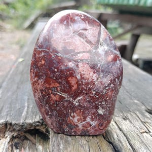 May include: A polished, oval-shaped stone in shades of deep red, maroon, and white. The stone has a smooth, glossy surface with natural patterns and veining. The stone is displayed on a weathered wooden surface.