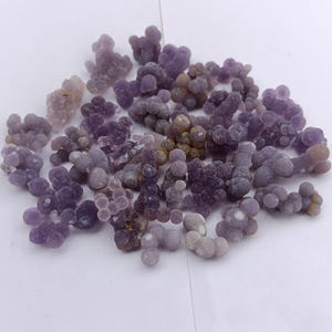 May include: A selection of grape agate gemstones in shades of purple and lavender. The stones have a clustered, bubbly appearance, similar to bunches of grapes. The gemstones are presented on a white surface.