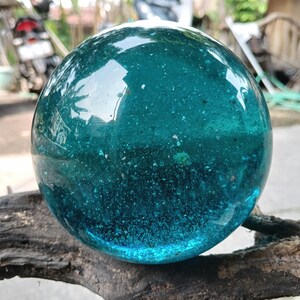 Könnte beinhalten: Eine große, klare, teal-farbene Glaskugel. Die Kugel hat eine leicht strukturierte Oberfläche und liegt auf einem Stück dunklem, verwittertem Holz. Das Glas reflektiert die Umgebung und erzeugt einen schimmernden Effekt.