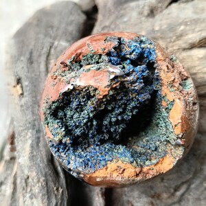 Puede incluir: Una piedra pulida y redonda con un exterior rugoso y texturizado. La capa exterior es de color marrón rojizo, mientras que el interior revela una cavidad llena de vibrantes formaciones cristalinas azules y verdes. La piedra es una geoda natural.