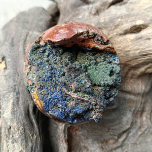 Puede incluir: Primer plano de un espécimen mineral pulido y redondo. La parte superior es de color marrón rojizo, mientras que la mitad inferior muestra una mezcla de tonos azules, verdes y marrones. El mineral tiene una superficie rugosa y texturizada, con estructuras cristalinas visibles.