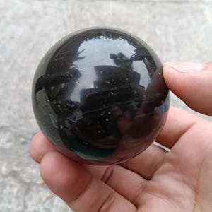 May include: A polished, spherical stone with a deep black, reflective surface. The smooth, rounded object features subtle, tiny white speckles embedded within the dark material, creating a glossy appearance that reflects the surrounding environment.