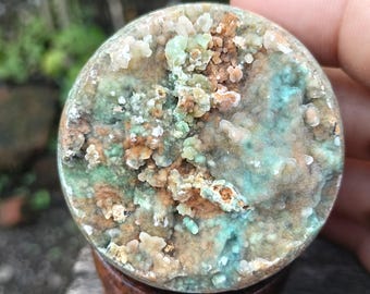 Puede incluir: Una piedra redonda y pulida con una superficie texturizada única. La piedra presenta una mezcla de colores, incluyendo tonos de verde, marrón y azul. La piedra está montada sobre una base de madera, mostrando su belleza natural.