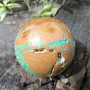 May include: A spherical, polished stone with a brown and green color scheme. The stone has a cracked appearance with veins of bright green running through the brown surface. The stone is resting on a weathered wooden surface.