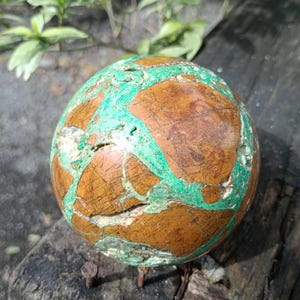 May include: A spherical decorative stone with a brown and green pattern. The stone has a polished surface with a network of green veins running across a brown base. The stone is sitting on a wooden surface.