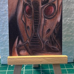 May include: A detailed drawing of a robotic figure with red eyes, displayed on a small wooden easel. The artwork features a brown and black color palette, with intricate mechanical details. The easel is made of light-colored wood.