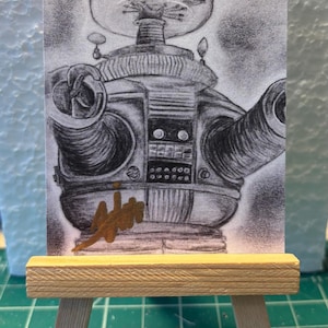 May include: A pencil sketch of a retro robot, possibly from a science fiction movie, is displayed on a small wooden easel. The robot has a round head, a cylindrical body, and two large, round arms. The artwork is in grayscale.