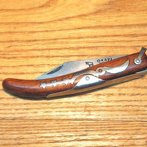 May include: A folding pocket knife with a curved wooden handle featuring silver-colored metal inlays in the shape of three stars and a crescent moon. The metal blade is partially visible and engraved with the word OKAPI and a small animal icon.