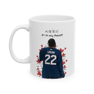 Puede incluir: Una taza de cerámica blanca con el gráfico de un jugador de hockey que viste una camiseta azul oscuro con el nombre LOGAN y el número 22 en la espalda. El diseño incluye corazones rojos y estrellas negras, con el texto #1 ON THE ICE #1 in my heart.