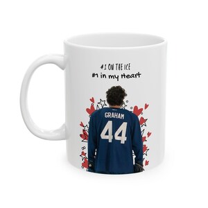 Puede incluir: Una taza de cerámica blanca con el gráfico de un jugador de hockey que viste una camiseta azul oscuro con el nombre GRAHAM y el número 44 en la espalda. El diseño incluye corazones rojos y estrellas negras, con el texto #1 ON THE ICE #1 in my heart.